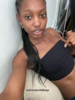 A person of color wearing a black bandeau top is positioned slightly off-center, looking towards the camera. The person has their hair styled with multiple braids and gold hoop earrings. The background is minimally visible, with a wall and what appears to be a clothing rack. The lighting is neutral, and the composition is a close-up, focusing on the upper body.