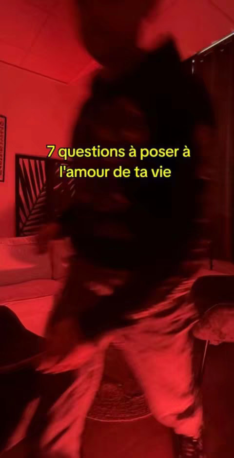 The image is dimly lit with a red hue. In the foreground, a person, blurry in the frame, is dancing or moving. The text overlay reads: '7 questions à poser à l'amour de ta vie' (7 questions to ask the love of your life). The background contains hints of a room setting.