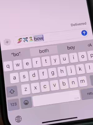 An iPhone screen displaying a text message interface. The keyboard is visible, with the user typing the word "bow." Above the keyboard, a text input field contains the word "bow," preceded by several colorful emojis. Suggestion words like "bo," "both," and "boy" are displayed above the keyboard, indicating options to complete the word or phrase. The overall color scheme is a gradient of light purples.