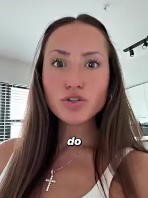 A young woman with long brown hair is centered in the frame, looking directly at the camera with wide eyes and a neutral expression. She is wearing a white tank top and a necklace with a cross pendant. The background is slightly blurred, with glimpses of a modern interior, visible lighting fixtures, and a window with vertical blinds to the left. The composition is a close-up, focusing on the woman’s face.