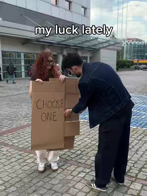 A street scene with two individuals interacting with a cardboard sign. A woman with red curly hair stands holding a sign that reads 'CHOOSE ONE'. A man, positioned on the right side of the frame, appears to be adjusting or interacting with the sign. The background shows a building entrance and pedestrian area.