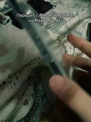 The image shows a hand holding a clear phone case over a patterned surface, possibly a bedspread or fabric. The phone is in the case and is partially visible, though the screen is not prominent. The hand dominates the foreground, and the patterned background provides context. The lighting appears to be natural, and the color palette is muted.