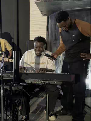 Two men are captured in a performance setting. One, seated, is playing a keyboard and smiling. The other, standing to the seated man's right, is holding a microphone and appears to be speaking or singing. The lighting is dim with a dark background.
