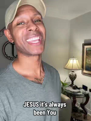 A Black man wearing a gray v-neck shirt and a beige cap is smiling widely at the camera from a medium shot. The background includes a lamp, a painting, and a small table.