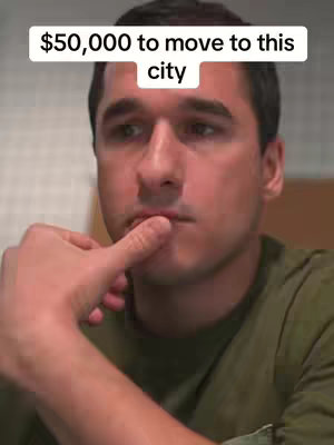 A man with short dark hair and a green shirt is looking off to the side, touching his lip with his index finger in a thoughtful pose. Above his head, there is white text that reads '$50,000 to move to this city.'
