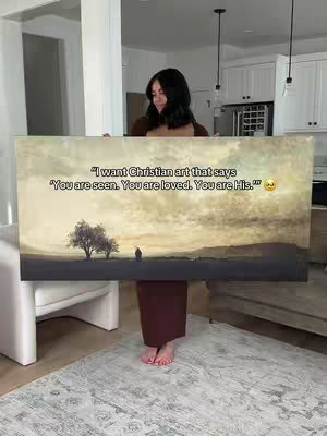 A woman stands in a well-lit room, holding a large rectangular canvas. The canvas displays a scenic landscape with a sky filled with clouds, trees, and a person in the distance. The woman wears a brown top and skirt, and her bare feet are visible.