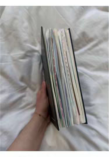 A human arm holding a half-open notebook or journal, angled slightly upwards. The pages inside are visible. The background is a white, textured surface, likely a bedsheet, creating a soft, neutral backdrop for the journal.