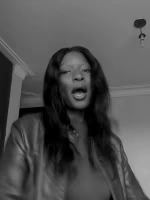 A black and white selfie-style video image of a woman with long dark hair, possibly in a home setting. She is looking directly at the camera with her mouth open. She appears to be in the middle of speaking or singing. The image is centered on her face and upper body, creating an immediate and personal connection.