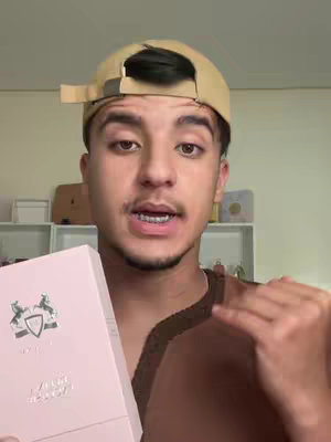 A young man in a beige baseball cap is holding a pink box with text and logo. He's looking at the camera while holding the box up, with an open mouth. The background includes a simple shelf. The overall shot is well-lit.