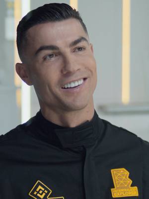 A medium shot of a man in a black uniform smiling and looking slightly to the side. The uniform has patches with logos. The background is a bright, possibly interior, setting. The overall composition suggests a promotional or advertisement-style image.