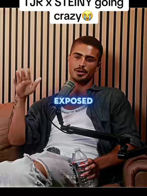 A man with a mustache sits indoors in a dimly lit room, with a microphone in front of him and a water bottle in his right hand. The backdrop appears to be vertical wooden slats, and a text overlay is positioned over the center of the image.