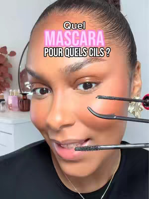 A close-up shot of a woman with dark skin, looking slightly to the right, holding mascara wands, as if testing or demonstrating them. She is wearing a dark top, with her hair pulled back. The backdrop includes a vanity setup.