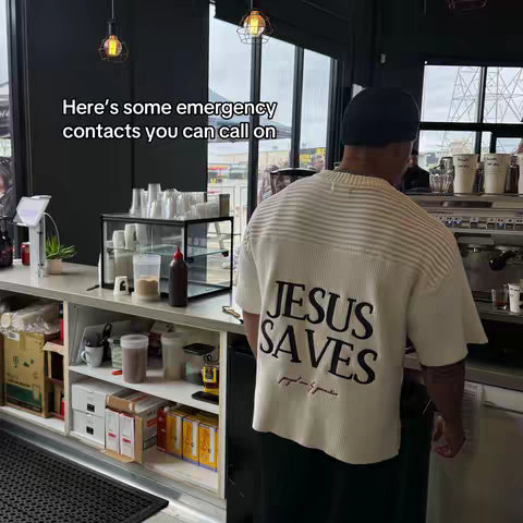 A man wearing a white t-shirt with 'JESUS SAVES' printed on the back is standing in a cafe setting. The image is from behind the man, obscuring his face, with the focus on his shirt and the surrounding details of the cafe. The background reveals elements of a coffee shop, including counters, equipment, and windows.