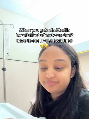 A selfie taken in a hospital room. A young woman with dark hair and a black shirt is looking down and to the right with a thoughtful expression. Above her head are two emoji of smiling faces and burger. In the background is an IV drip stand and a hospital room.