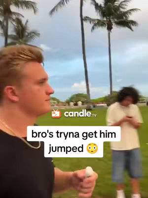A selfie-style video shows a young man in a black shirt on the left side of the screen, holding something white in his hand, while another person in a t-shirt and shorts is in the background, standing on the grass. Palm trees and a cloudy sky form the backdrop, indicating an outdoor setting.