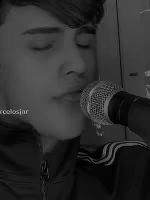 A black and white image shows a close-up of a person singing into a microphone. The person is a young man with dark hair. The microphone is positioned close to his mouth. He is wearing a dark jacket with white stripes on the sleeves. The color of the image is desaturated.