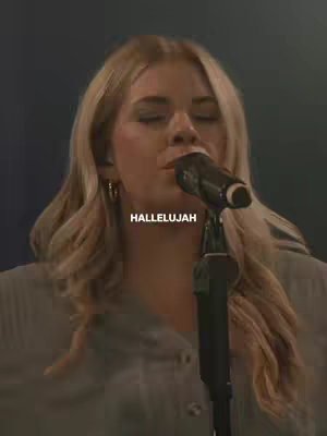 A medium shot of a woman with long blonde hair, lit with soft, warm light. She is holding a microphone up to her mouth, eyes closed, as though she is singing. The text overlay "HALLELUJAH" is visible beneath her.