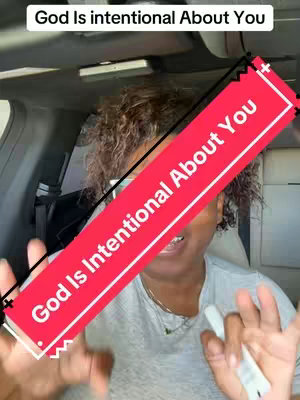 A woman in a car, smiling and looking directly at the camera. She's holding one finger over her lips, a red banner across her chest with the white text 'God Is Intentional About You'. The background is the interior of a vehicle.