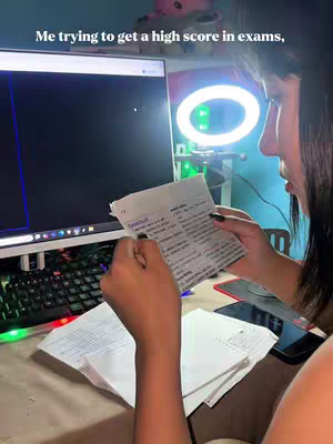 A young woman is shown in profile, studying at a desk with a computer and a ring light. She is looking down at a paper with notes on it.