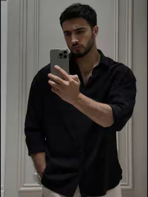 A man with dark hair and a beard is taking a selfie in front of a mirror. He is wearing a black button-down shirt and white pants. The background appears to be a white wall and doorframe.