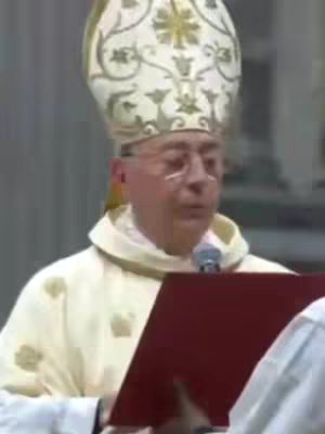 The image shows a man in religious attire. He is wearing a white headdress with gold trim and a gold garment. He is holding a red book and speaking into a microphone. The background is blurred, giving focus to the man.