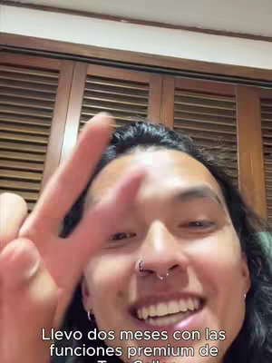 A selfie-style video with the creator taking up most of the frame. The creator is a young person with dark hair, a nose ring, and earrings, smiling and holding up a peace sign with two fingers. The background shows wooden paneling, likely a wall or door. The foreground includes text describing the premium features of Truecaller.