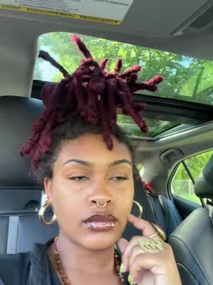 A selfie-style video shot from inside a car. A woman with dark skin and vibrant, burgundy-colored locs is the primary focus. She wears large gold hoop earrings, a nose ring, and has her hand near her chin, with a gold ring visible on her finger. The car's interior features a black seat and a glimpse of the outside scenery through the windshield and sunroof.