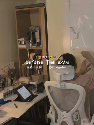 A well-lit interior shot shows a desk area with a wooden shelf unit in the background. On the desk, a laptop is open, and the chair faces away from the camera. The text overlay says "before the exam."