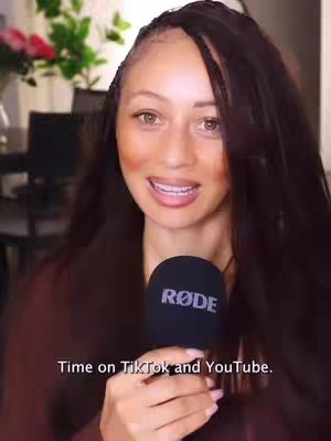 A woman with long dark hair holds a black microphone close to her face, smiling slightly. The background is blurred, showing hints of an interior setting with floral decorations.