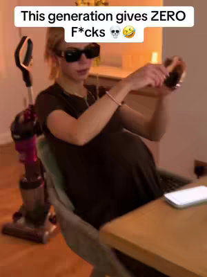 A person, wearing black sunglasses and a dark top, sits in a chair holding a glass while looking at the phone. A vacuum cleaner is next to the chair. The background is a room with warm lighting.