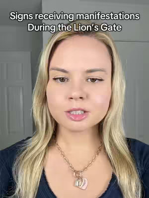 A woman with long blonde hair is centered, looking directly at the camera. Above her, the text overlay reads, "Signs receiving manifestations During the Lion’s Gate." She's wearing a navy blue top and a gold necklace with a charm. The background is a light-colored wall.