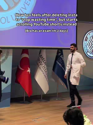 A man in a white lab coat and black pants is walking across a stage. Behind him are several flags and a screen with text on it. The flags are Turkish and have the logo of the Medipol University.