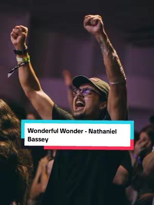 A man with his arms raised in the air with clenched fists, eyes closed and mouth open in an expression of joy and excitement. The setting is indoors, possibly a concert or event. The image is framed with a title bar overlayed at the bottom with the artist name and song title.