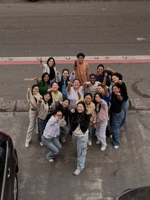 An elevated, high-angle shot capturing a group of approximately 18 individuals, predominantly appearing to be young adults. They are arranged in a roughly circular formation on a paved surface, likely a sidewalk or parking area, with a road and a vehicle visible in the background. The group is unified in raising their fists in a gesture of solidarity or excitement. The lighting appears natural, and the scene conveys a sense of vibrancy and community.