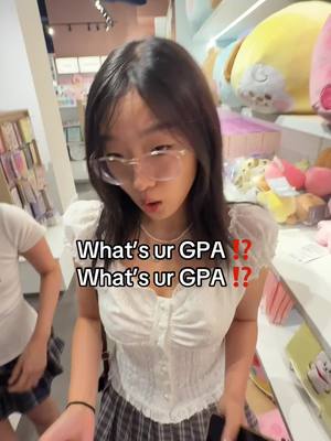 A young woman wearing glasses looks into the camera with a surprised expression. Text overlays read "What's ur GPA ?!" The background is a brightly lit store with merchandise displayed on shelves.
