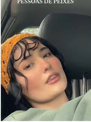 A selfie-style shot featuring a person facing the camera, sitting inside a car. The individual wears a mustard-colored crocheted hat and a light green top. The background is blurred but suggests the inside of a vehicle.