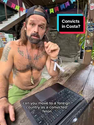 A man with long hair, a backward baseball cap, and tattoos sits at a wooden table with a laptop. A text overlay describes the video's topic. The background includes foliage and a stone statue.