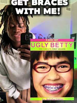 A person with dark hair and dreadlocks is holding a phone to take a selfie. The background includes a mirror and a partial view of a room. To the right, there's a DVD case of "Ugly Betty" tv series. Above the creator, a text overlay reads "GET BRACES WITH ME!"