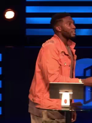 A man in an orange jacket stands at a podium-like table, speaking in front of a blue-lit background with vertical stripes.
