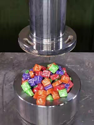 An overhead shot features an industrial press descending onto a container filled with colorful candies. The press is a large, cylindrical metal piece with a flat, circular base, positioned above a matching container. The container is filled with wrapped square candies in various colors, creating a vibrant display. The composition is simple yet striking, with a focus on the contrast between mechanical and colorful elements.