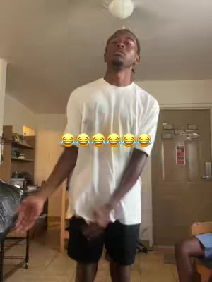 A young Black man is captured in a medium shot, posing in a well-lit indoor space. He is wearing a white t-shirt and dark shorts. The background is slightly blurred, revealing a door and other room elements. His expression is animated and a series of emoji stickers.
