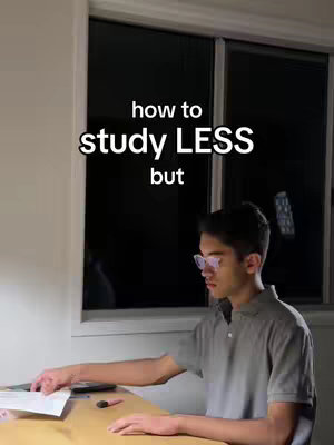 A well-lit interior shot featuring a young man at a desk near a window. The man, wearing glasses and a grey polo shirt, is leaning over the desk, interacting with papers. Text in the upper portion of the frame reads, 'how to study LESS but'. The background consists of a large dark window and a portion of the room wall. The setting conveys a calm, focused atmosphere.