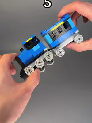 A close-up shot of a person's hands holding a Lego train. The train is made of blue, yellow, gray, and white Lego bricks. The number "5" is in white and is located in the upper part of the image.