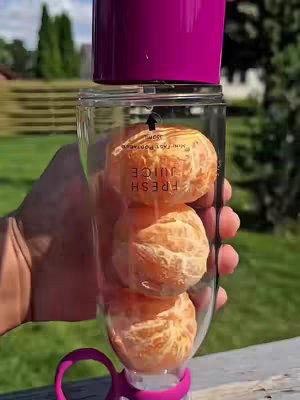 A hand holds a clear plastic cup or bottle, which has three peeled oranges inside, with a pink colored top. The background is a green yard and trees.