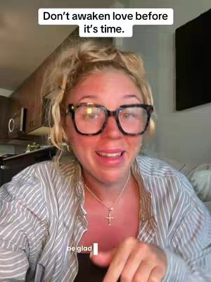 A woman with blonde hair, wearing large black-framed glasses, and a striped shirt is speaking to the camera. Text overlayed on the image reads, 'Don't awaken love before it's time.' and 'be glad'. The background shows a kitchen setting.
