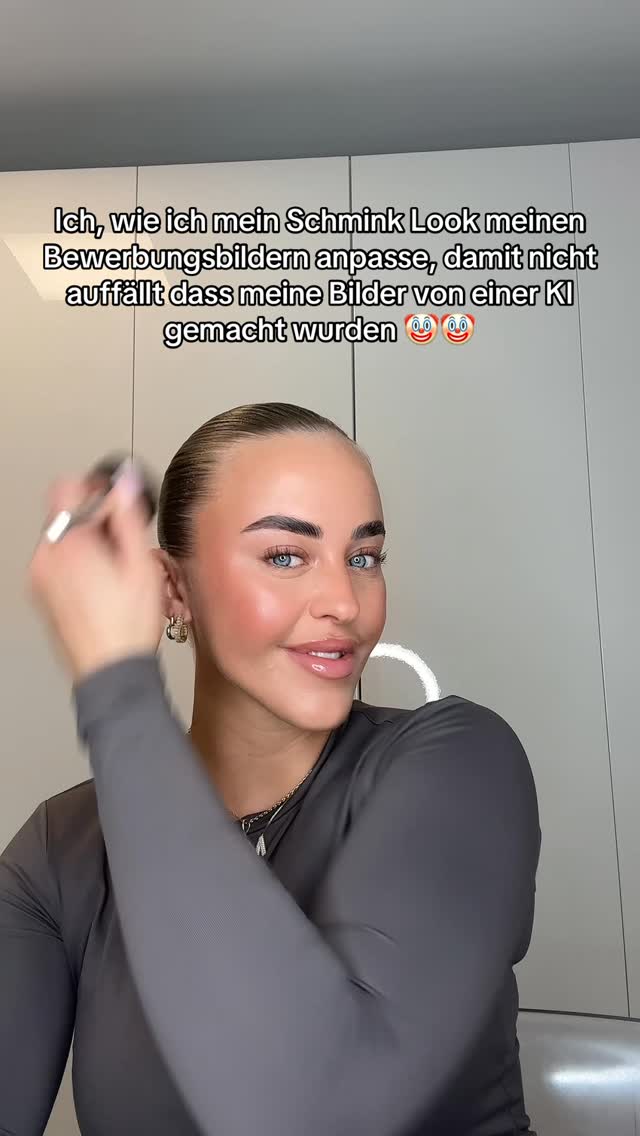 A woman is applying makeup, with a close-up shot of her face. She has fair skin, blue eyes, and is wearing a dark grey top. The background is a plain, off-white wall, and text is overlaid explaining she is adapting her makeup to her job-application photos so it is not obvious that they were created with AI. Her hair is pulled back in a sleek, low bun and she is holding a makeup brush to her cheek.