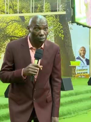 A bald man wearing a maroon suit and a pink shirt is holding a microphone, looking towards the camera with a slightly concerned expression. He is standing indoors in front of a backdrop that appears to be a stage.