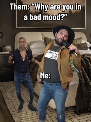 The image shows a meme with two men in a bedroom setting. On the right, a man holds a microphone and appears to be singing, with text overlayed that says 'ME'. On the left, another man is dancing. The upper portion contains text written in a meme format, 'Them: 'Why are you in a bad mood?''. The room features a bed with a patterned headboard and a dark tapestry.