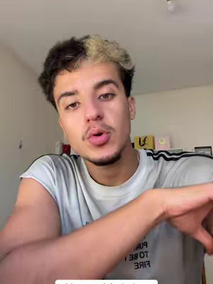 A young man with short, dark hair and a section of blonde hair, is centered in the frame, looking towards the camera with a slight pout. He wears a light grey t-shirt. The background comprises a plain wall, a hanging light fixture, and various items on a shelf, including boxes and decorative elements.