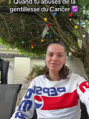 A woman is seen smiling at the camera in a shot filmed outdoors. She's wearing a white sweatshirt featuring a red and blue design on the front. Overhead, there is a tree with colorful lights strung across its branches. The background shows a house with a glass door.